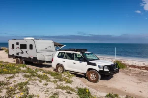 Landscape aerial view of 4WD and modern caravan parked adjacent to a sparkling sunny beach.