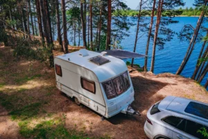 Aerial view of Caravan RV parked on the beach under a trees facing blue sea, lake or river. Family traveling with motorhome. Travelers on an active family vacation.